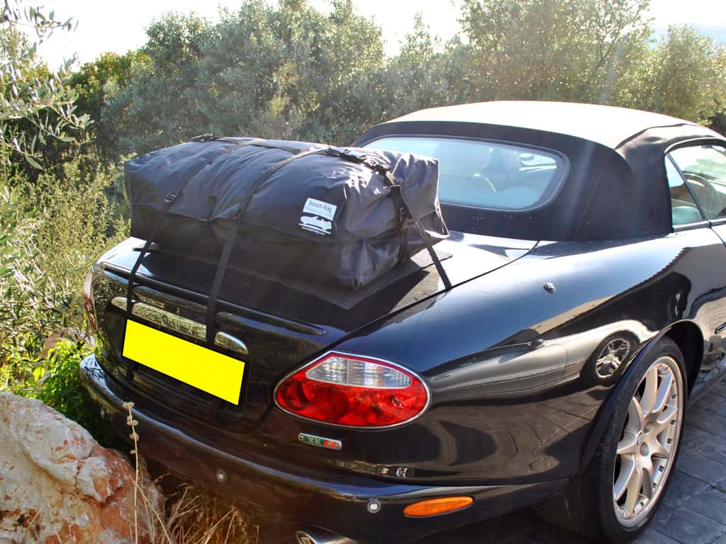 black jaguar xkr convertible with the hood up and a boot-bag vacation luggage rack fitted next to a wooded area.