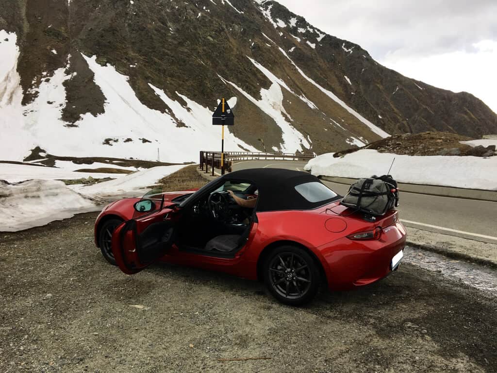 red mazda mx5 mk4 with a luggage rack fitted with a grey holdall on it on a snowy mountain pass