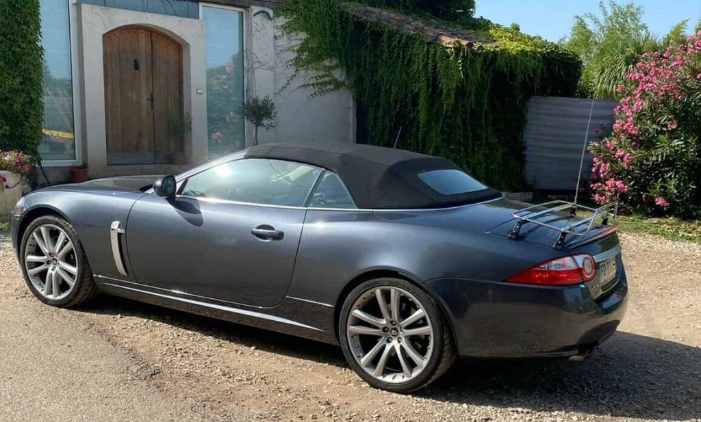 dark grey jaguar xk convertible with a stainless steel luggage rack fitted outside a cottage on a sunny day
