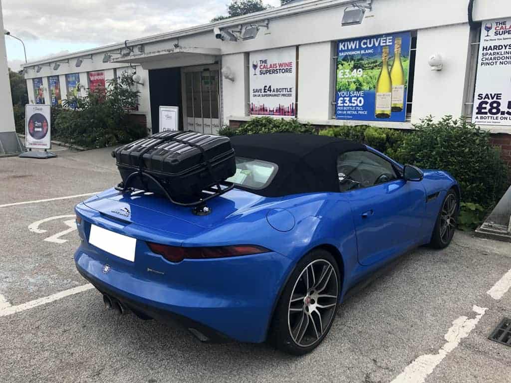 blue jaguar f type convertible with a black luggage rack fitted carrying a suitcase outside a wine shop