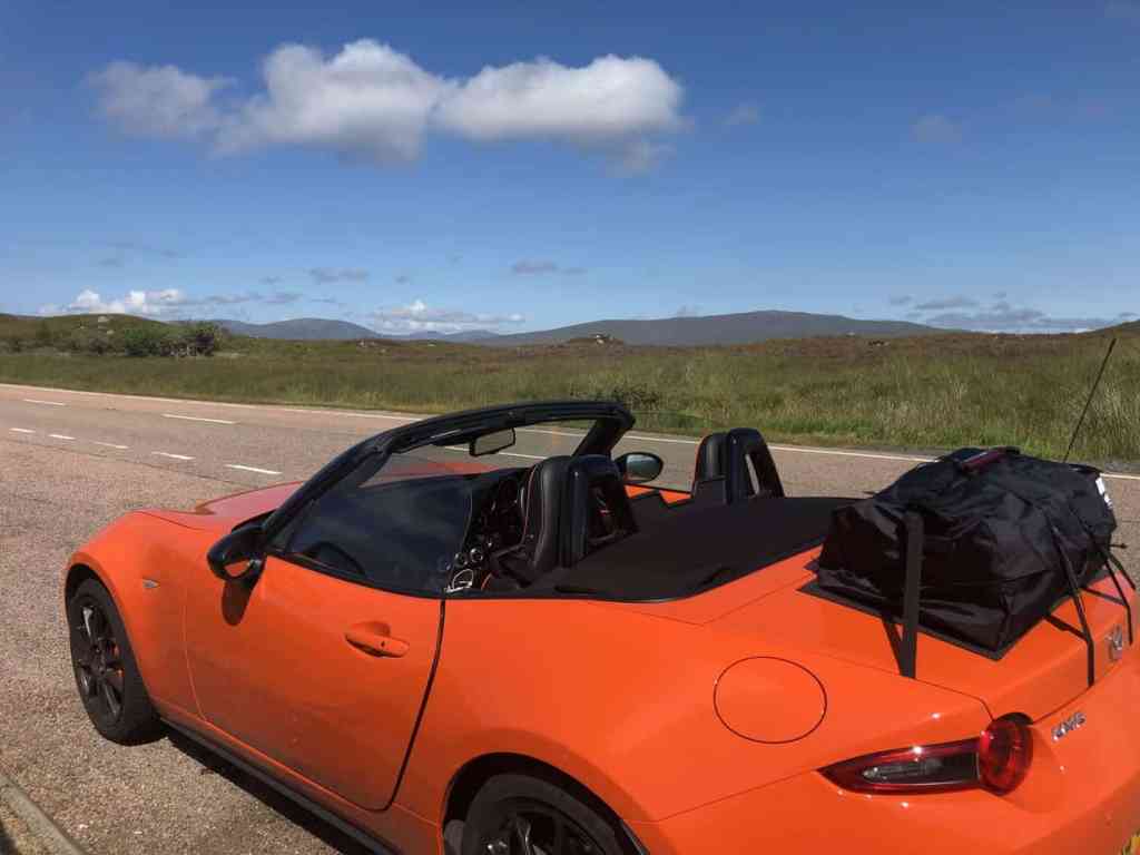 Orange mazda mx5 mk4 with a boot-bag luggage rack fitted on a scottish country road with the roof down overlooking hills