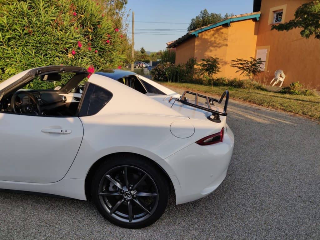 white mazda mx5 rf with a black luggage rack fitted on. rural road next to a cottage on a sunny evening 