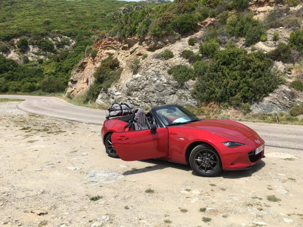 red mazda mx5 mk4 on a Corsica mountain road roof down with a revo-rack luggage rack fitted carrying a grey bag