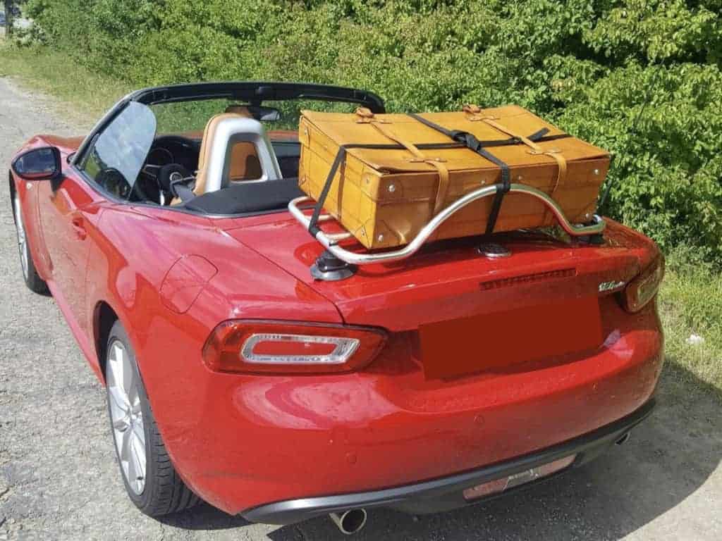red fiat 124 spider roof down next to a hedge with a stainless steel luggage rack fitted carrying large beige case
