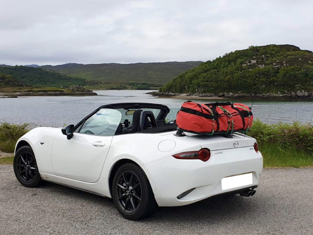 white mazda mx5 nd mk4 with a revo-rack black luggage rack fitted carrying a red holdall parked opposite a lake roof down