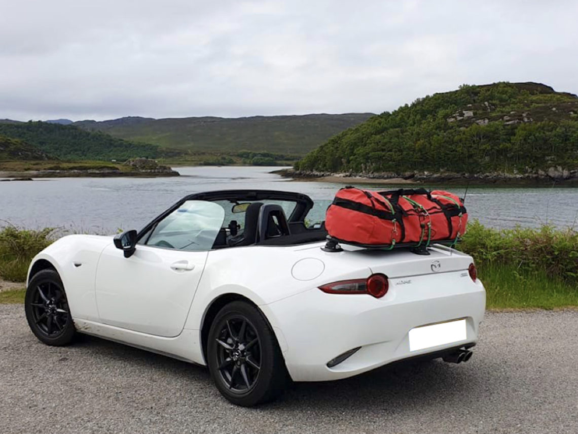 white mazda mx5 nd next to a lake with a luggage rack fitted carrying a red bag