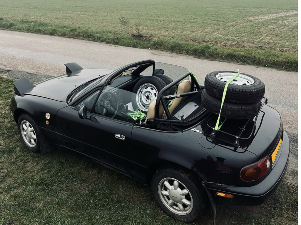 black Mazda mx5 na mk1 with a luggage rack fitted carrying two spare wheels photographed from above next to a field 